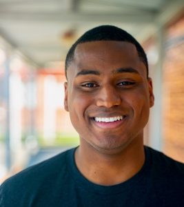 A young man with short hair and a black T-shirt smiles warmly while standing in a bright, covered outdoor walkway. The background is softly blurred.
