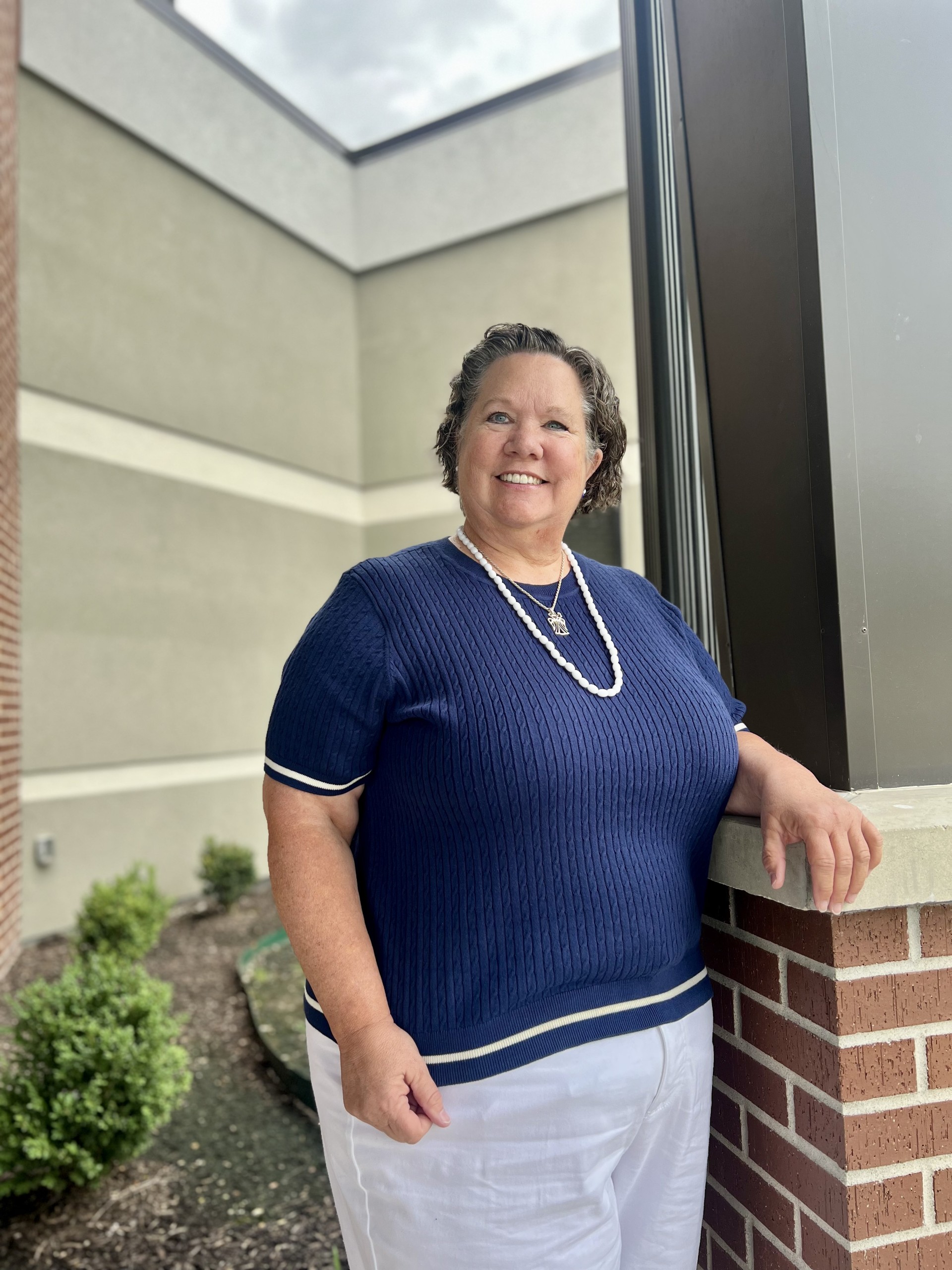 A smiling woman with short, curly hair wears a navy blue top, white pants, and a pearl necklace, standing outdoors by a brick and concrete building with shrubs in the background.