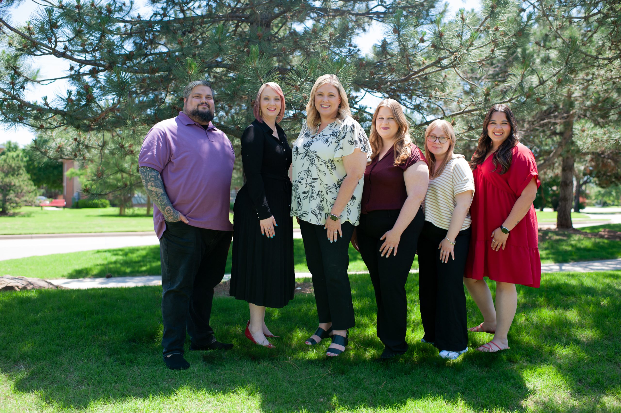 Six people, five women and one man, stand together outdoors on grass in front of leafy trees, smiling at the camera on a sunny day. They are dressed in casual and semi-formal clothing.