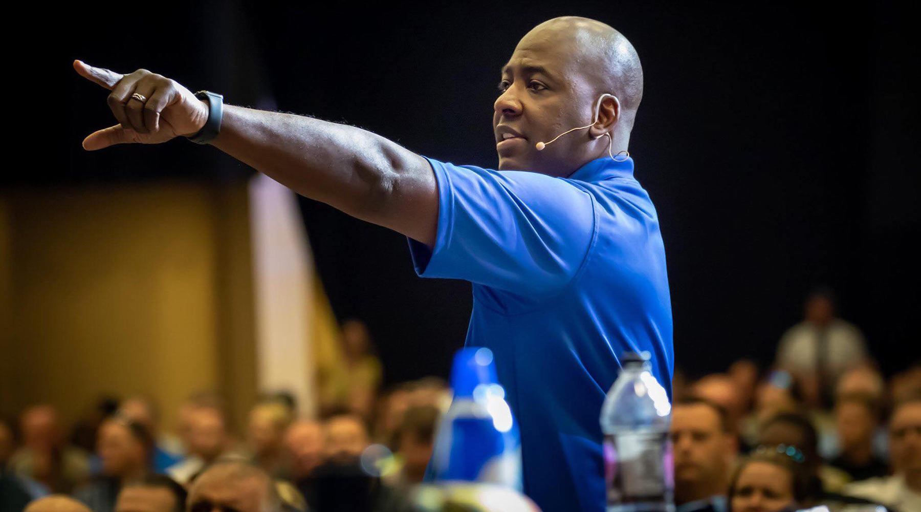 A man wearing a blue polo shirt and a headset microphone gestures while speaking to a seated audience in a large indoor venue. A water bottle and blue cup are visible in the foreground.