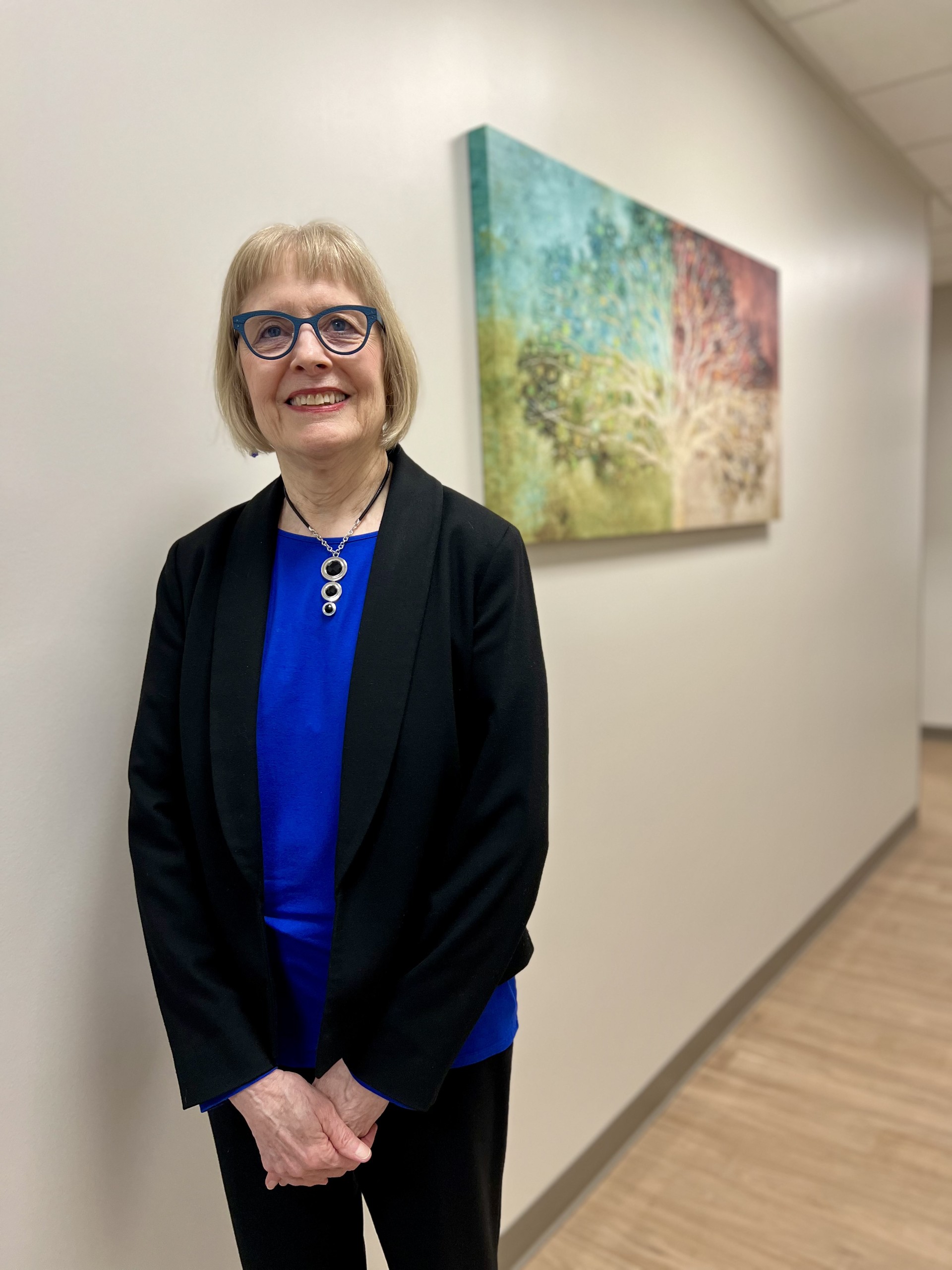 Smiling woman with short blond hair, glasses, and a black blazer stands in a hallway with wood floors. Colorful abstract tree artwork hangs on the wall behind her.